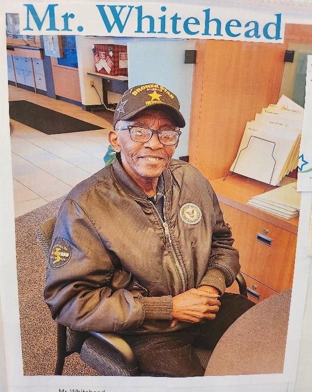 An older Black man sitting in a chair and smiling. He is wearing a cap and jacket that identify him as a veteran and Bronze Star Hero