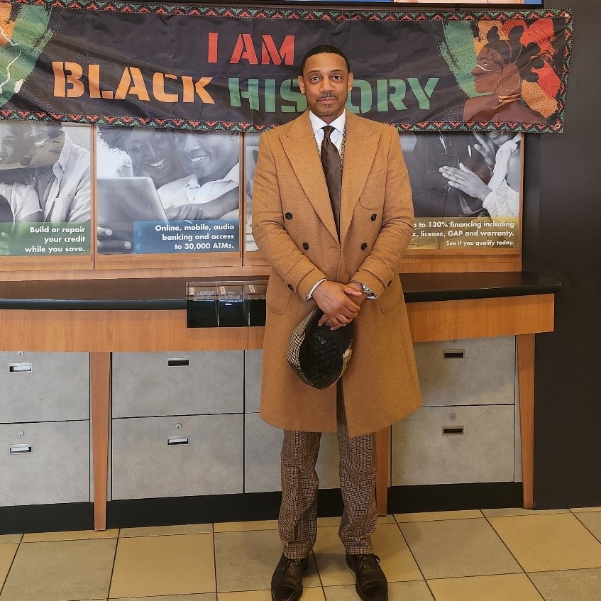 A well-dressed Black man in a credit union lobby standing beneath a banner that says 