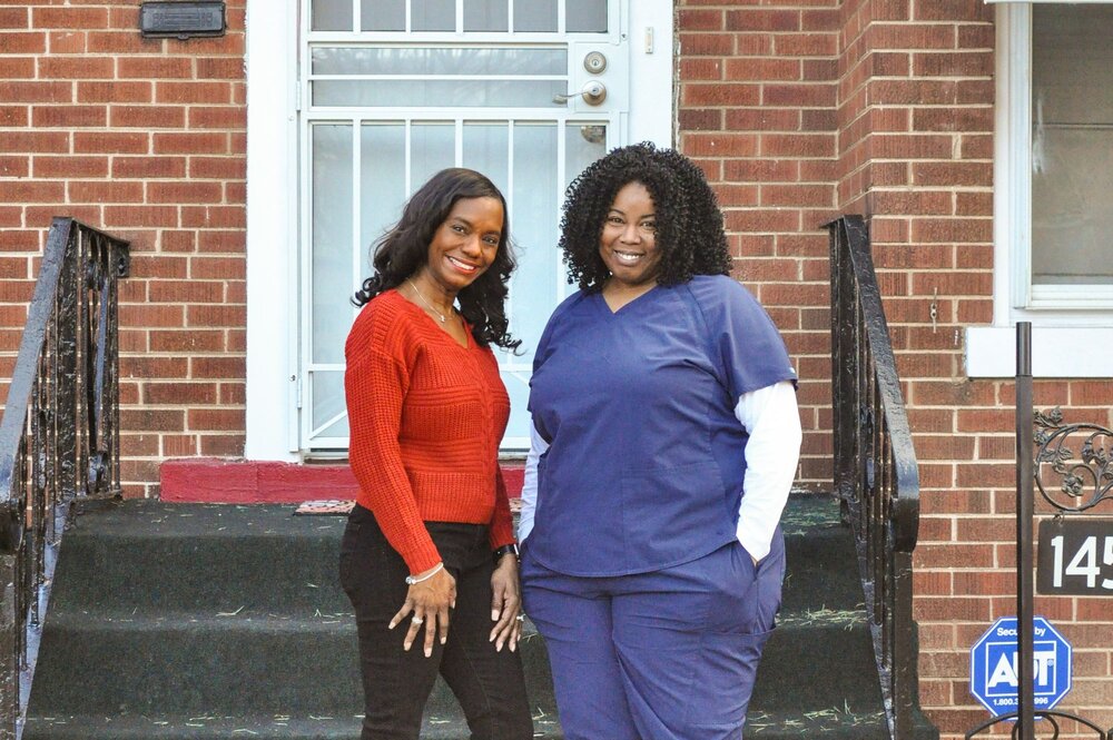 two smiling Black women standing in front of a city home