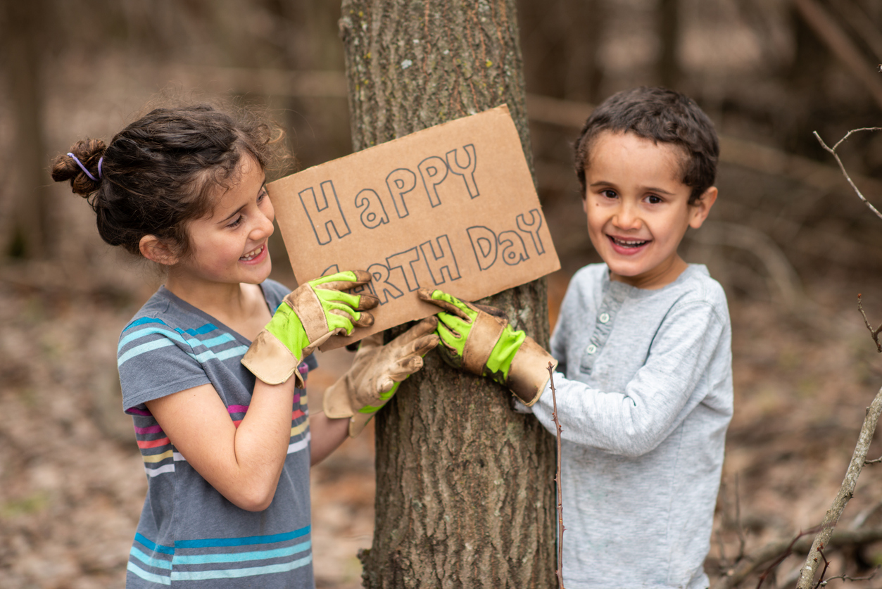 Two smiling children standing by a tree holding a cardboard sign that says Happy Earth Day