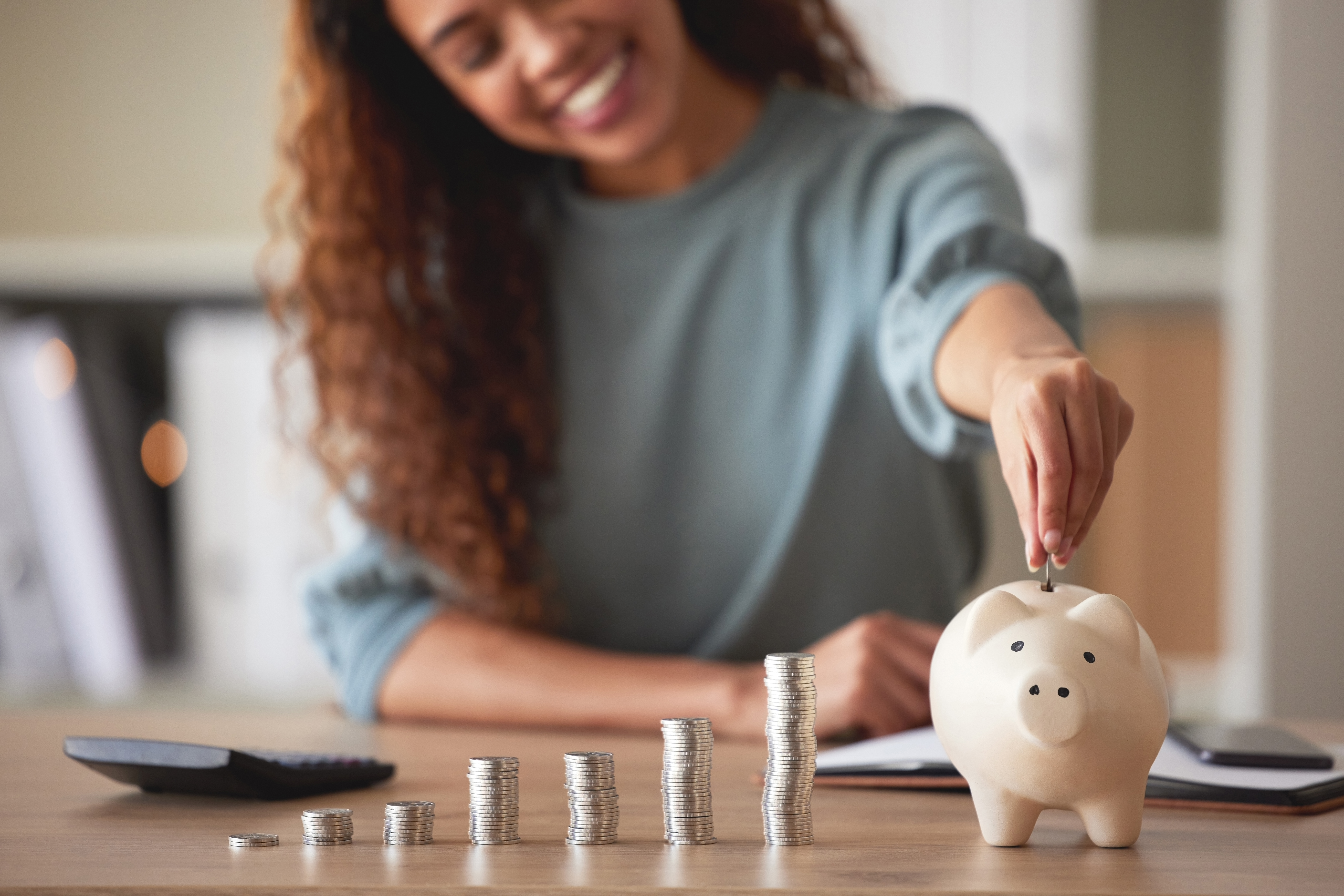 image of a young person putting coins into a piggybank at home with several stacks of coins next to the piggybank